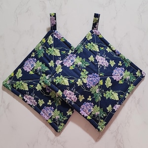 May include: Two navy blue square pot holders with a floral pattern of purple hydrangeas and green leaves. Each pot holder has a small fabric loop for hanging. The edges are trimmed with a green border.