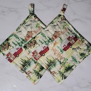 May include: Two quilted pot holders with a camping theme. The cream-colored fabric features a pattern of red and white camper trailers, bears, deer, and trees. Each pot holder has a hanging loop.