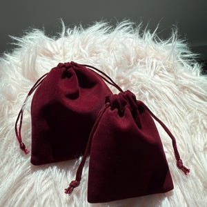 May include: Two deep burgundy velvet drawstring pouches of different sizes. Each pouch features a matching fabric cord closure with knotted ends, designed to cinch the top securely. The soft, matte texture of the velvet fabric is visible.