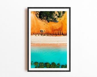 May include: A framed aerial photograph featuring a vibrant landscape with a bright turquoise ocean, a strip of white sand beach, and a contrasting orange-red cliffside topped with dark green trees, all enclosed in a simple black frame.