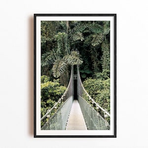 May include: A framed vertical wall art print featuring a narrow metal suspension bridge extending into a dense, dark green tropical rainforest. The bridge has a light-colored walkway and wire mesh railings, surrounded by lush palm leaves and foliage.