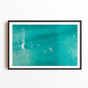 Puede incluir: Vista aérea de surfistas en agua turquesa. La imagen, enmarcada en negro, captura a varios surfistas sobre sus tablas, con estelas blancas de algunos. La escena se desarrolla en un día soleado y brillante.
