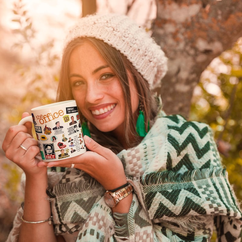 May include: A white ceramic mug with a colourful design featuring characters and quotes from the TV show "The Office". The mug is being held by a person wearing a beige knitted hat and a green and white patterned scarf.