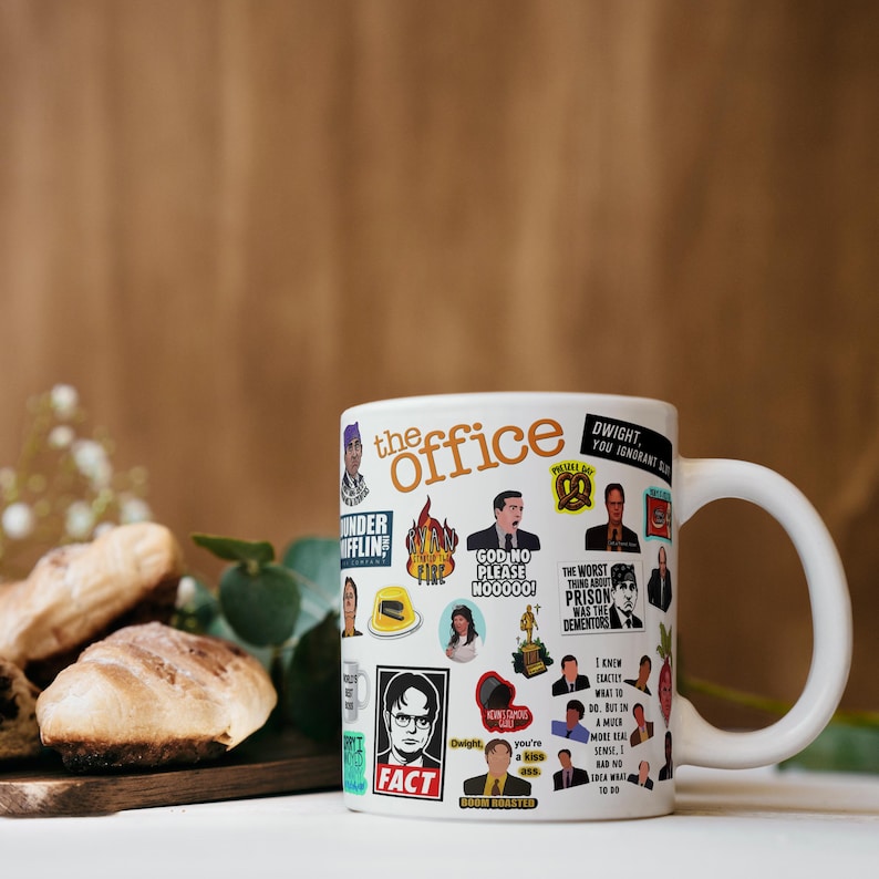 May include: White ceramic mug with a colourful design featuring characters and quotes from the TV show "The Office". The mug has a black handle and the words "The Office" are printed in orange.