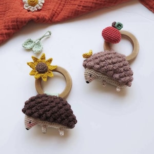 Op de afbeelding: Twee gehaakte babyrammelaars in de vorm van egels met houten ringen. Eén heeft een zonnebloem, de andere een rode appel. Extra gehaakte bloemen en bladeren zijn verspreid.