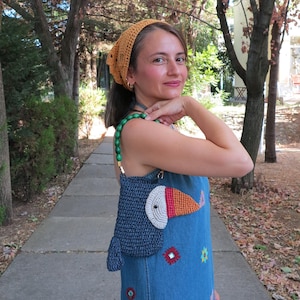 May include: A blue crochet handbag with a toucan design, featuring a white, orange, and red beak. The bag has a green beaded strap and is held by a person wearing a denim dress with floral embroidery and a yellow headscarf.