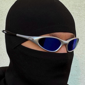 May include: Silver-framed sunglasses with blue lenses are worn over a black balaclava. The sunglasses have a sleek, modern design. The balaclava covers the head and neck, leaving only the eyes visible.