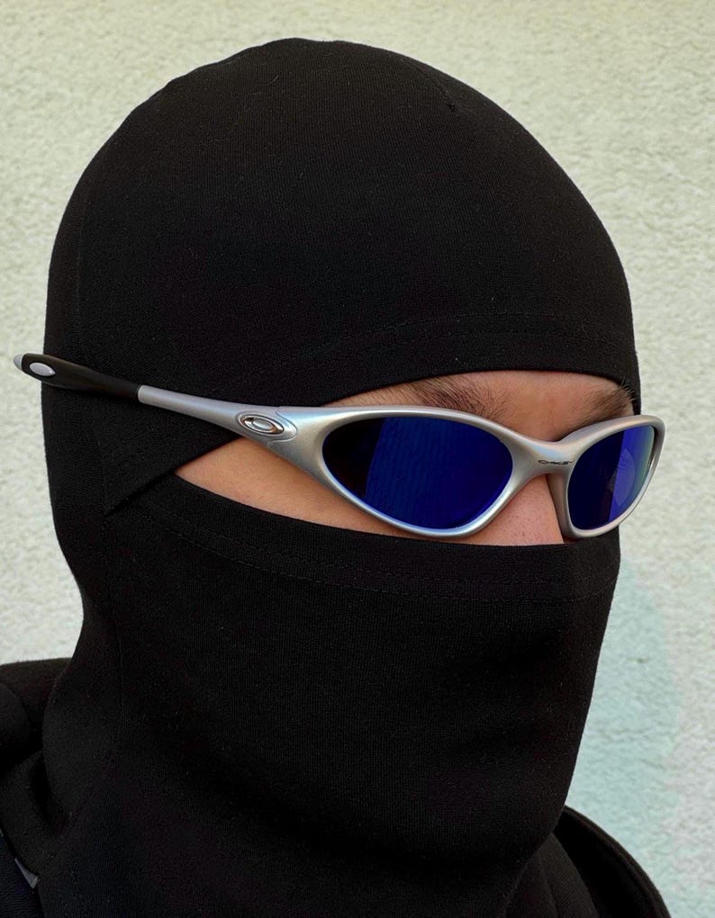 May include: Silver-framed sunglasses with blue lenses are worn over a black balaclava. The sunglasses have a sleek, modern design. The balaclava covers the head and neck, leaving only the eyes visible.