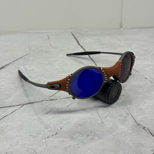 May include: Sunglasses with a brown leather frame stitched around the lenses. The lenses are blue and the arms are a dark gray color. The sunglasses are resting on a black cylindrical object.