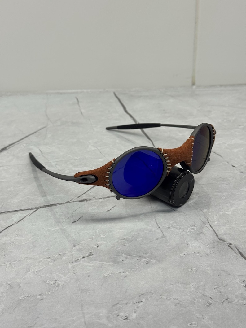 May include: Sunglasses with a brown leather frame stitched around the lenses. The lenses are blue and the arms are a dark gray color. The sunglasses are resting on a black cylindrical object.