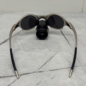May include: Silver-framed sunglasses with dark lenses and a black nose bridge. The sunglasses have a sleek, modern design with curved temples and black ear socks. The lenses are oval-shaped and appear to be polarized.
