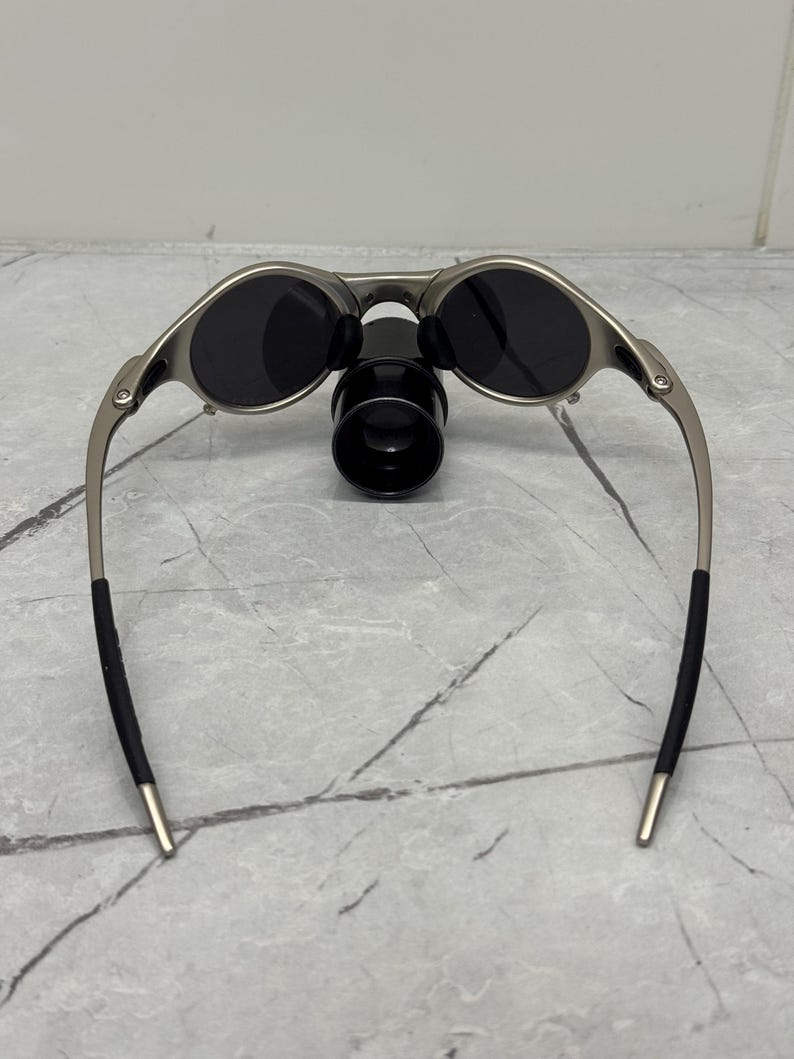 May include: Silver-framed sunglasses with dark lenses and a black nose bridge. The sunglasses have a sleek, modern design with curved temples and black ear socks. The lenses are oval-shaped and appear to be polarized.