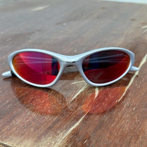 May include: Silver-framed sunglasses with red lenses. The sunglasses have a curved design and the brand name "Oakley" is visible on the frame. The lenses reflect the surrounding environment. The sunglasses are resting on a wooden surface.