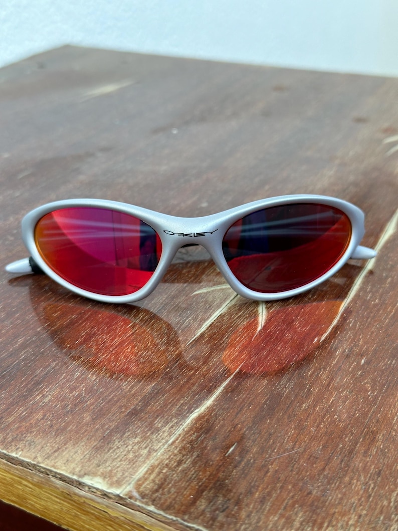 May include: Silver-framed sunglasses with red lenses. The sunglasses have a curved design and the brand name "Oakley" is visible on the frame. The lenses reflect the surrounding environment. The sunglasses are resting on a wooden surface.
