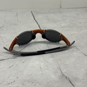 May include: Sunglasses with a brown leather bridge and temples, and dark gray lenses. The frame is a dark gray color. The sunglasses are resting on a light gray surface.