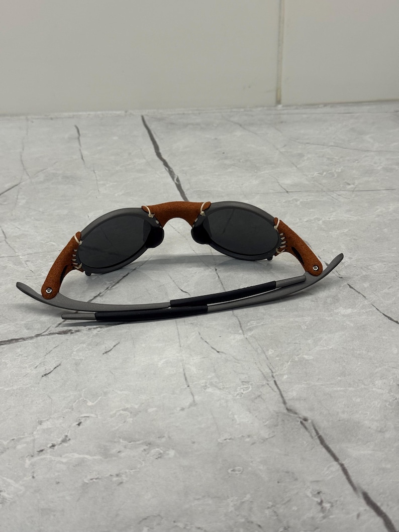 May include: Sunglasses with a brown leather bridge and temples, and dark gray lenses. The frame is a dark gray color. The sunglasses are resting on a light gray surface.
