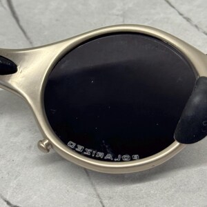 May include: Close-up of a pair of sunglasses with a metallic frame and dark lenses. The lenses are round and the frame has a matte finish. The word "POLARIZED" is printed on the frame.
