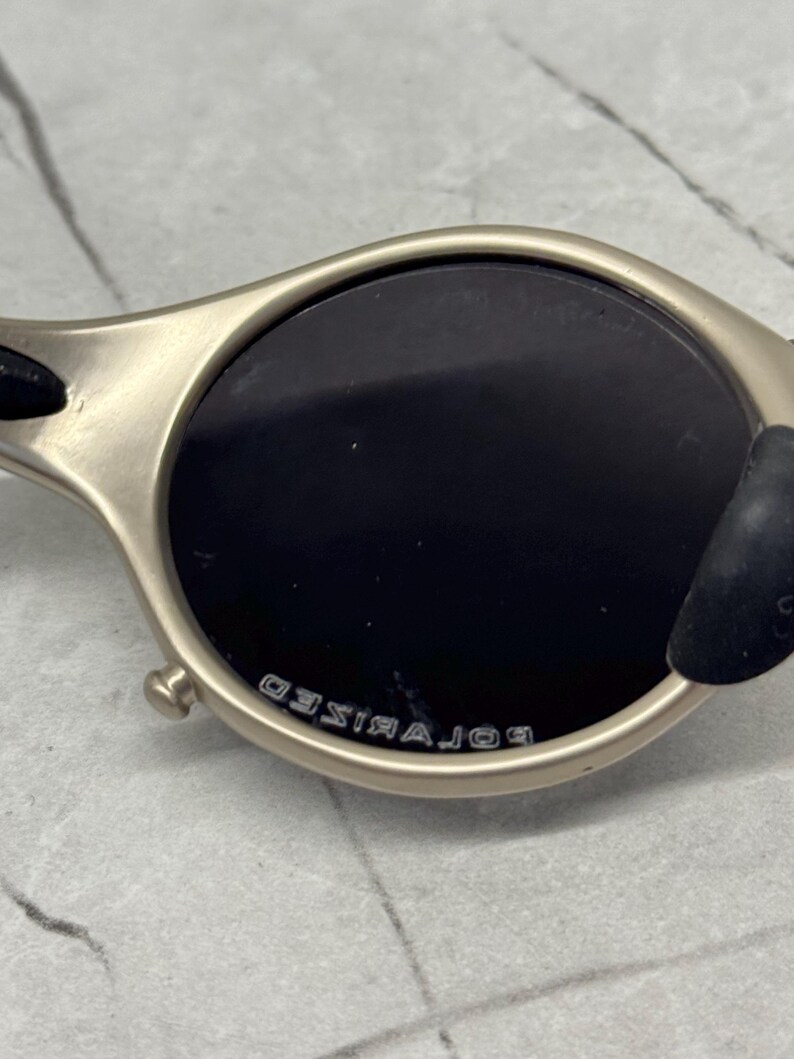 May include: Close-up of a pair of sunglasses with a metallic frame and dark lenses. The lenses are round and the frame has a matte finish. The word "POLARIZED" is printed on the frame.