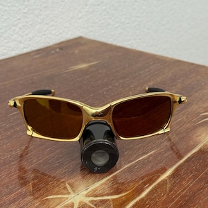 May include: Gold-framed sunglasses with brown lenses and a black attachment. The sunglasses have a rectangular shape and are resting on a wooden surface. The attachment has the text "8x".