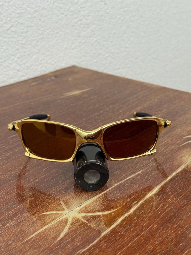 May include: Gold-framed sunglasses with brown lenses and a black attachment. The sunglasses have a rectangular shape and are resting on a wooden surface. The attachment has the text "8x".