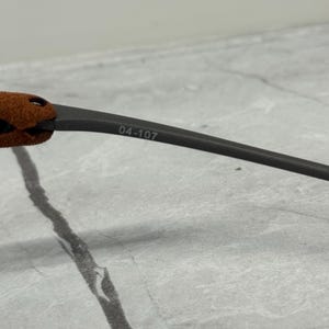 May include: Close-up of a dark gray eyeglass arm with the number "04-107" printed on it. The arm has a brown suede-like material near the hinge. The background is a light gray surface.