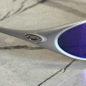 May include: Silver sunglasses with purple lenses. The sunglasses have a sleek, modern design with a metallic finish. The lenses are a vibrant purple colour, and the frame features a small logo near the temple.