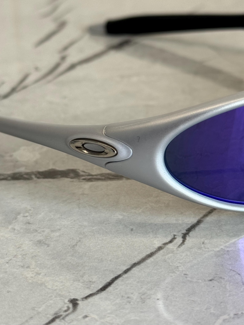 May include: Silver sunglasses with purple lenses. The sunglasses have a sleek, modern design with a metallic finish. The lenses are a vibrant purple colour, and the frame features a small logo near the temple.