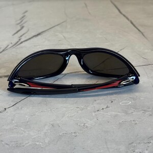 May include: Black sunglasses with dark lenses. The frames are black with red accents on the arms. The sunglasses are lying on a gray, textured surface.