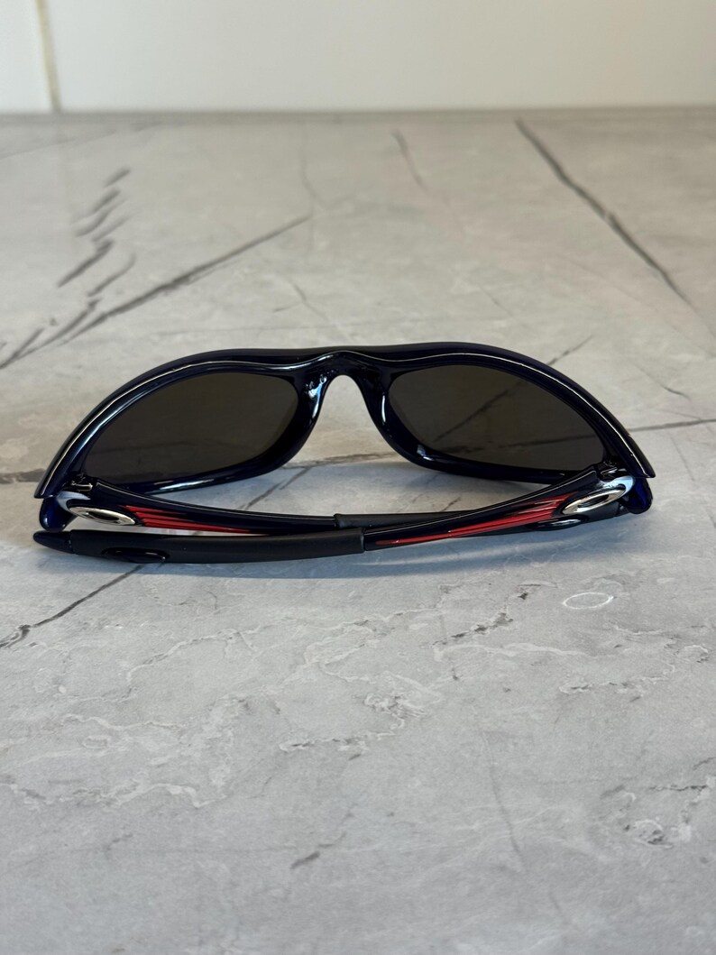 May include: Black sunglasses with dark lenses. The frames are black with red accents on the arms. The sunglasses are lying on a gray, textured surface.