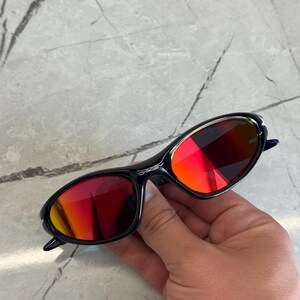 May include: Black sunglasses with red-tinted lenses. The sunglasses have a sleek, oval shape and a dark frame. The lenses reflect a gradient of red and orange hues. The brand name is visible on the frame.