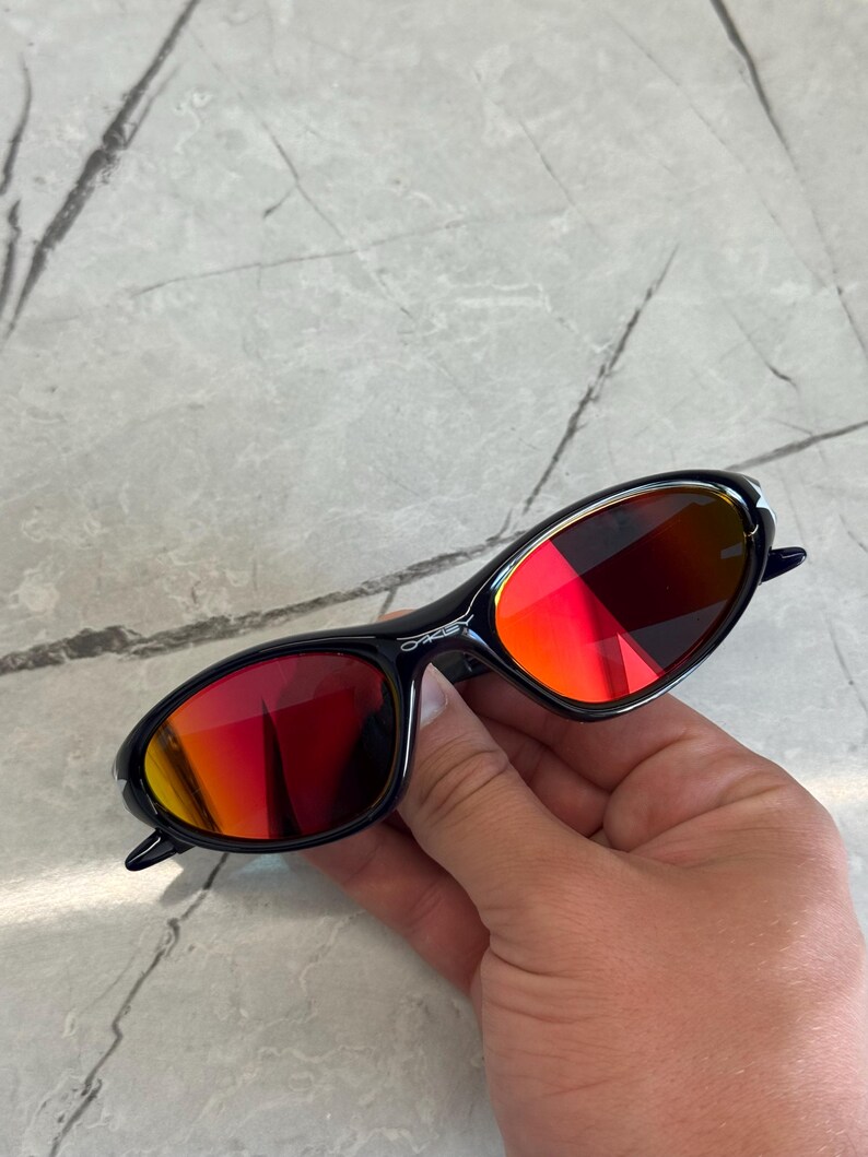 May include: Black sunglasses with red-tinted lenses. The sunglasses have a sleek, oval shape and a dark frame. The lenses reflect a gradient of red and orange hues. The brand name is visible on the frame.