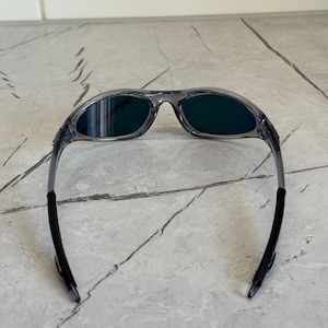K&ouml;nnte beinhalten: Grau gerahmte Sonnenbrille mit dunkelgr&uuml;nen Gl&auml;sern. Die Sonnenbrille hat ein sportliches Design mit einem gebogenen Rahmen und schwarzen Akzenten an den B&uuml;geln. Die Gl&auml;ser sind dunkelgr&uuml;n.