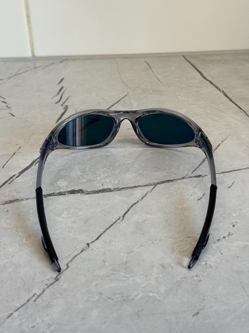 K&ouml;nnte beinhalten: Grau gerahmte Sonnenbrille mit dunkelgr&uuml;nen Gl&auml;sern. Die Sonnenbrille hat ein sportliches Design mit einem gebogenen Rahmen und schwarzen Akzenten an den B&uuml;geln. Die Gl&auml;ser sind dunkelgr&uuml;n.