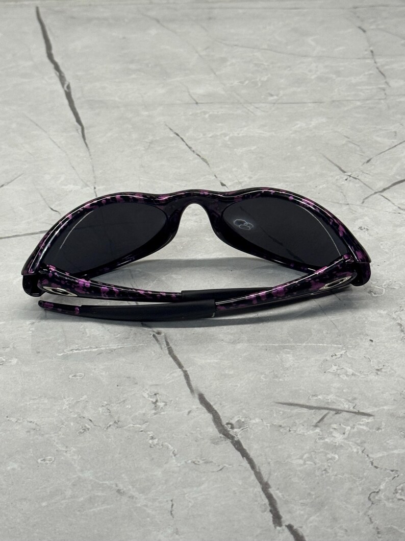 May include: Black sunglasses with a purple and black speckled pattern on the frames. The sunglasses have dark lenses and a sleek design. The arms are black and fold inwards.