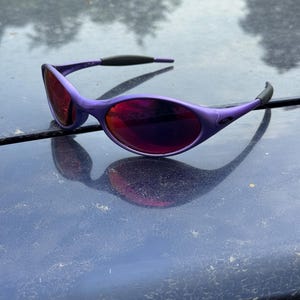 May include: Purple sunglasses with a curved frame and dark red lenses. The sunglasses have a sleek, modern design with black accents on the temples. The lenses reflect the surrounding environment.