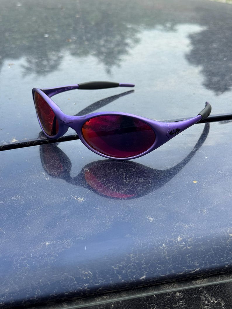 May include: Purple sunglasses with a curved frame and dark red lenses. The sunglasses have a sleek, modern design with black accents on the temples. The lenses reflect the surrounding environment.