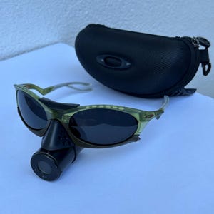 May include: Green and black sunglasses with a black case. The sunglasses have a unique design with a dark lens and a black attachment. The case is black and has a zipper closure. The sunglasses are on a white surface.