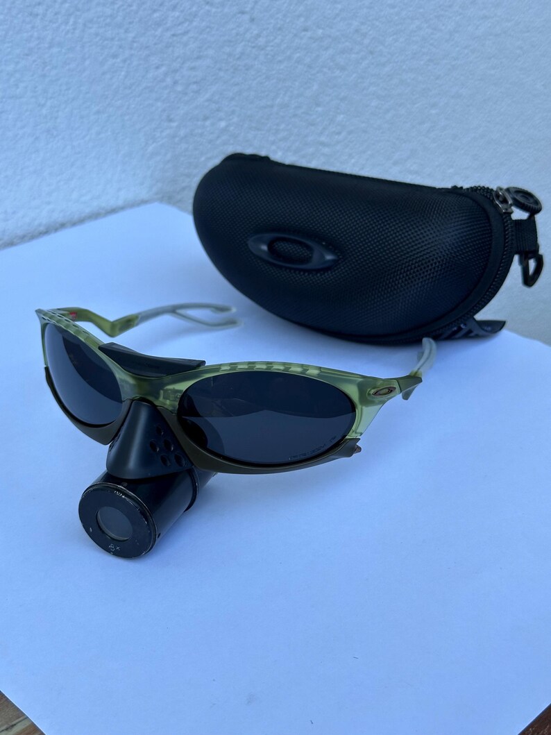 May include: Green and black sunglasses with a black case. The sunglasses have a unique design with a dark lens and a black attachment. The case is black and has a zipper closure. The sunglasses are on a white surface.