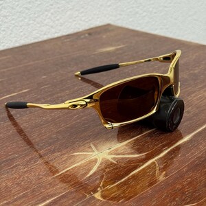 May include: Gold-framed sunglasses with brown lenses. The sunglasses have a sleek, modern design with black earpieces. The frame is a shiny gold color. The sunglasses are resting on a wooden surface.