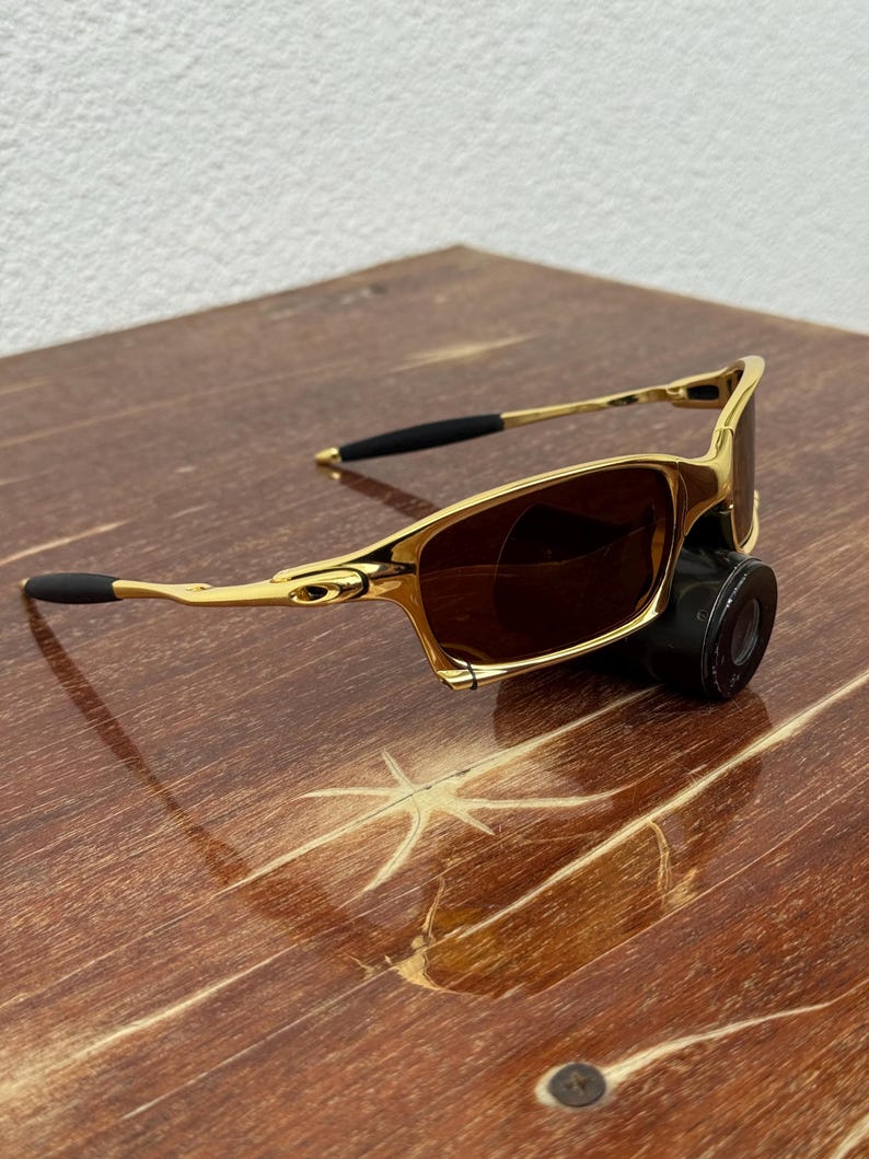 May include: Gold-framed sunglasses with brown lenses. The sunglasses have a sleek, modern design with black earpieces. The frame is a shiny gold color. The sunglasses are resting on a wooden surface.
