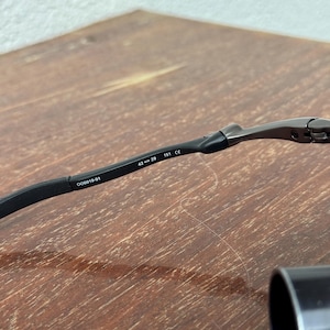 May include: Close-up of a black eyeglass frame with the text "006019-01" and measurements "42-29 151 CE". The frame is on a wooden surface.