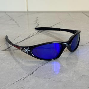 May include: Black sunglasses with blue lenses. The frames have a curved design and feature a star and logo detail on the temples. The lenses reflect a bright blue color.