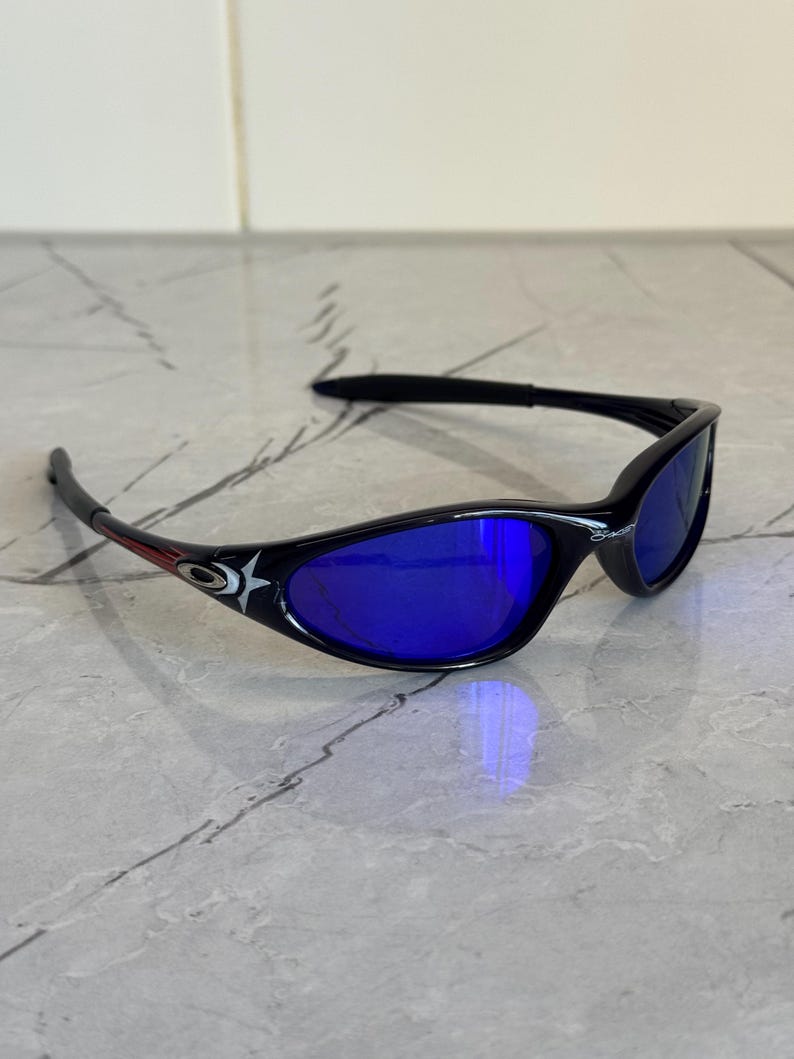 May include: Black sunglasses with blue lenses. The frames have a curved design and feature a star and logo detail on the temples. The lenses reflect a bright blue color.