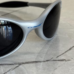 Puede incluir: Gafas de sol plateadas con lentes oscuras. Las gafas de sol tienen un dise&ntilde;o curvo y el nombre de la marca "Oakley" est&aacute; impreso en azul en la montura. Las gafas de sol se muestran sobre una superficie de m&aacute;rmol.