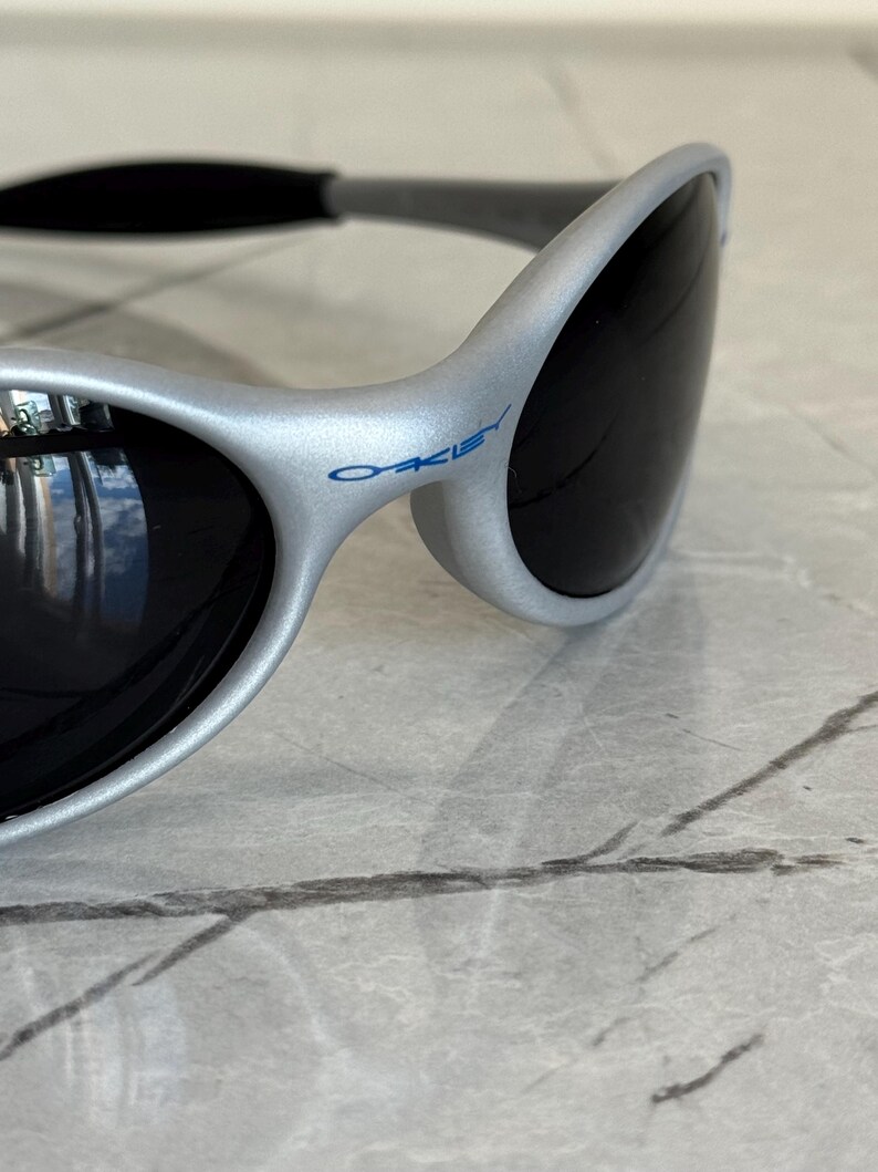 Puede incluir: Gafas de sol plateadas con lentes oscuras. Las gafas de sol tienen un dise&ntilde;o curvo y el nombre de la marca "Oakley" est&aacute; impreso en azul en la montura. Las gafas de sol se muestran sobre una superficie de m&aacute;rmol.