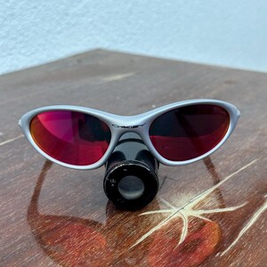 May include: Silver-framed Oakley sunglasses with red lenses. The sunglasses are resting on a dark brown wooden surface. The Oakley logo is visible on the frame.