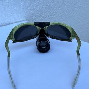 May include: Green-tinted sunglasses with dark lenses and a black bridge. The frames have white dot accents and silver-colored arms. A black, cylindrical attachment is centered between the lenses.