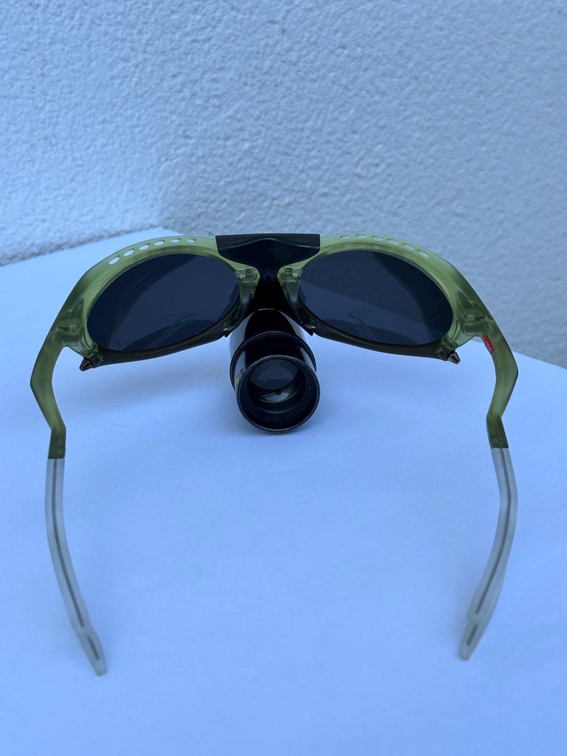 May include: Green-tinted sunglasses with dark lenses and a black bridge. The frames have white dot accents and silver-colored arms. A black, cylindrical attachment is centered between the lenses.