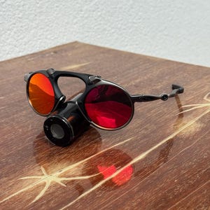 May include: Black and silver sunglasses with red lenses. The sunglasses have a unique design with a central cylindrical component and circular lenses. The lenses reflect light, and the frame is a dark color. The sunglasses are on a wooden surface.