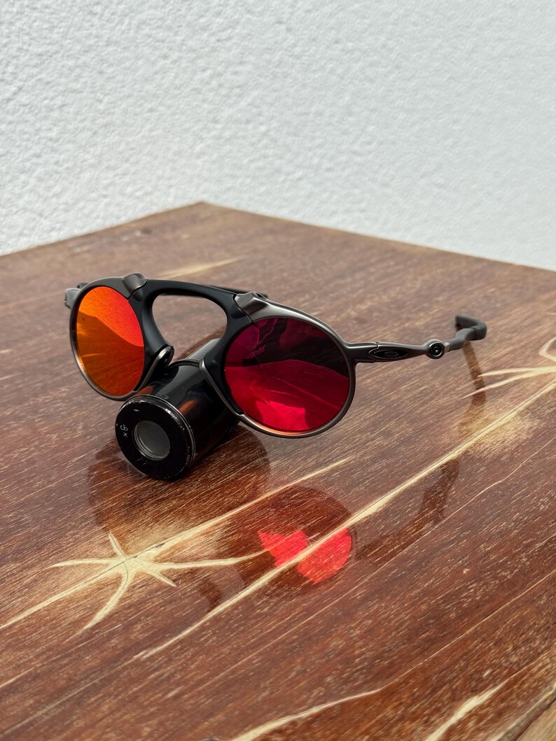 May include: Black and silver sunglasses with red lenses. The sunglasses have a unique design with a central cylindrical component and circular lenses. The lenses reflect light, and the frame is a dark color. The sunglasses are on a wooden surface.